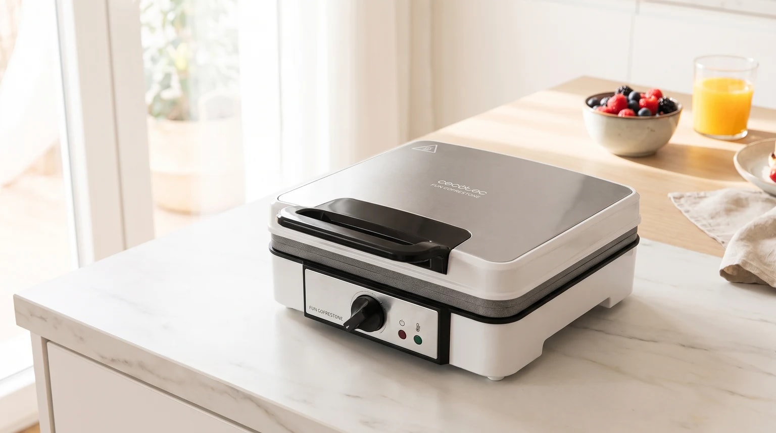 Uma máquina de fazer waffles branca e prateada numa bancada de cozinha, com uma taça de frutos vermelhos e sumo de laranja ao fundo.