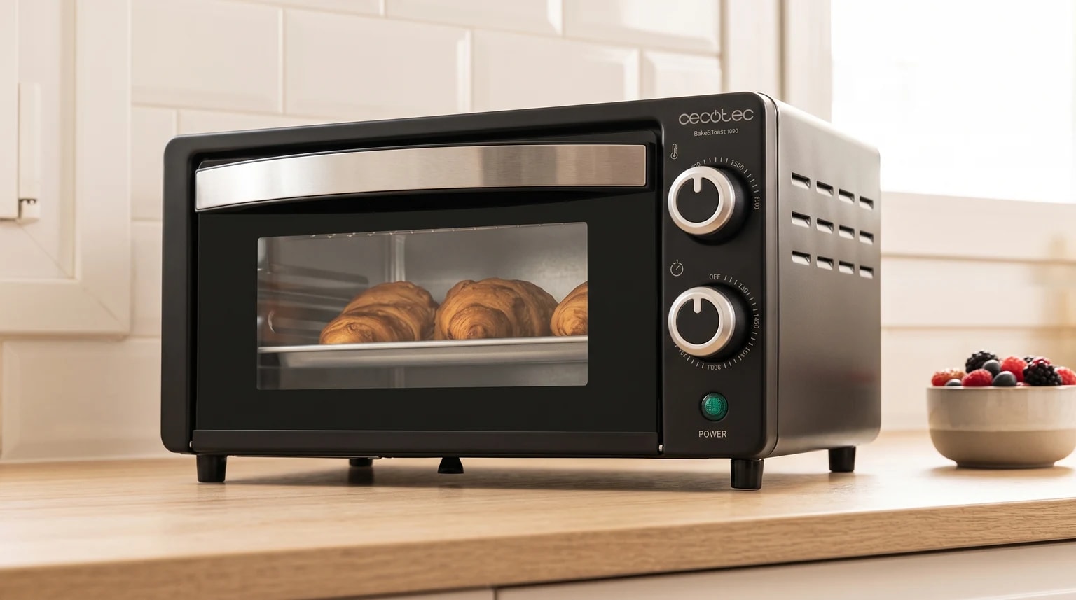 Mini forno elétrico Cecotec Bake&Toast 1090 preto sobre uma bancada de madeira, com três croissants a cozer e uma taça de frutos vermelhos ao lado.