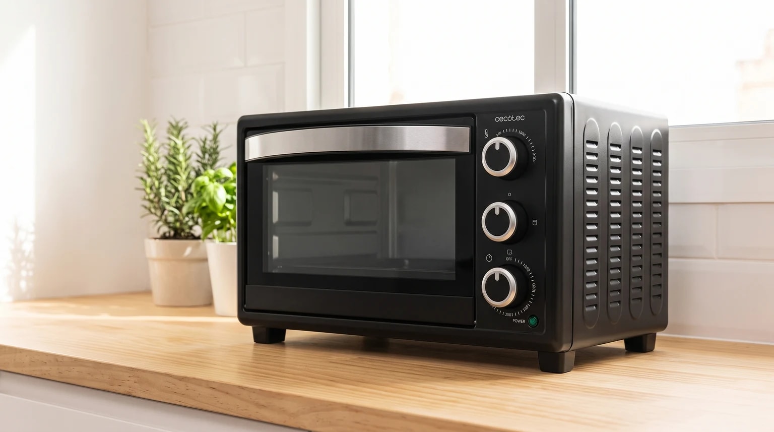Mini forno elétrico Cecotec Bake&Toast 2300 preto sobre uma bancada de madeira clara, com plantas e uma janela ao fundo.
