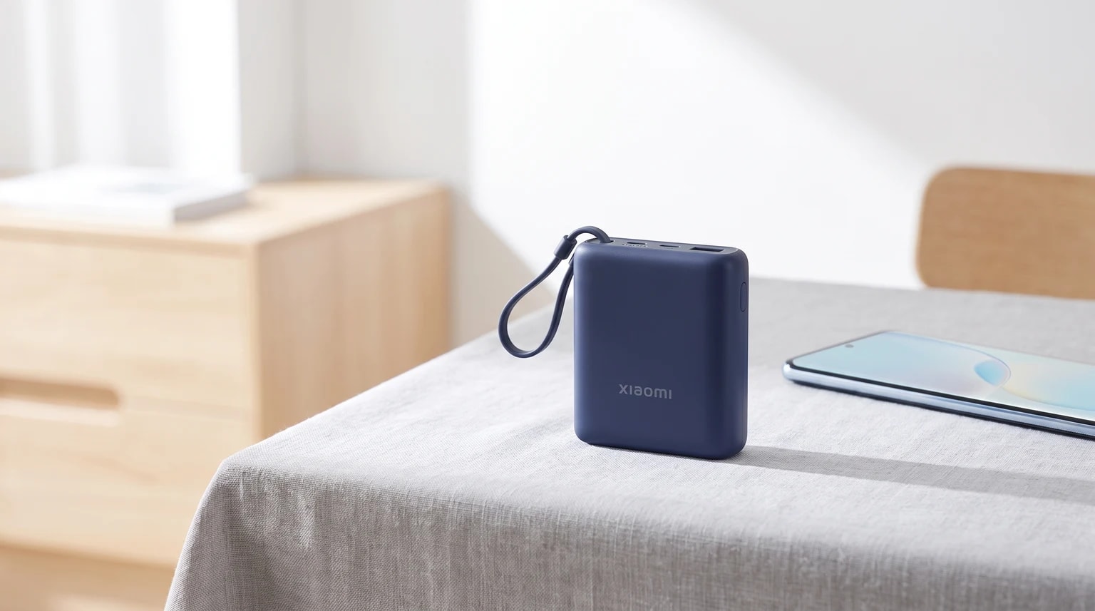 Uma powerbank compacta azul da Xiaomi, com um cabo integrado em forma de asa, sobre uma mesa com toalha de linho ao lado de um smartphone.
