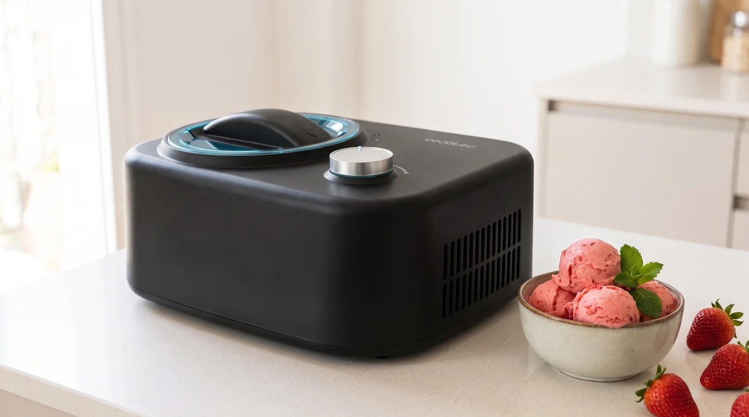 Máquina de fazer gelados Cecotec Gelacy 800 preta numa bancada de cozinha, ao lado de gelado de morango e morangos frescos.
