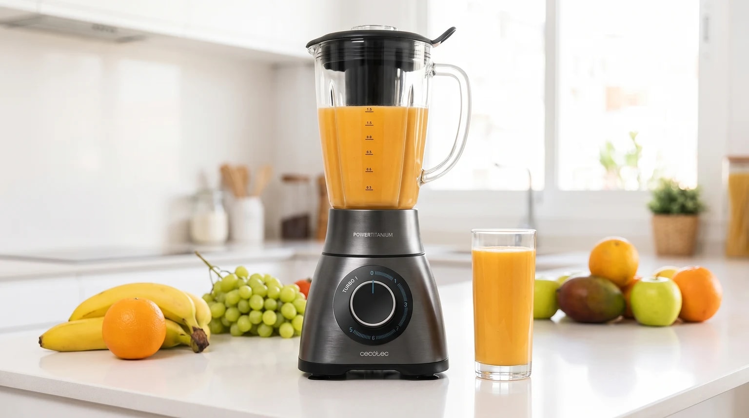 Liquidificador cinzento numa bancada de cozinha, com um batido de laranja no jarro e um copo servido ao lado, rodeado de várias frutas frescas.