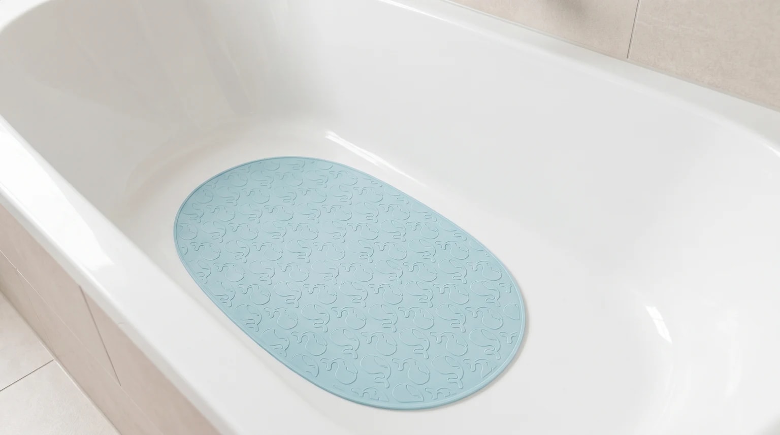 Tapete antiderrapante oval azul-claro com relevo de elefantes pousado no fundo seco de uma banheira branca e luminosa.