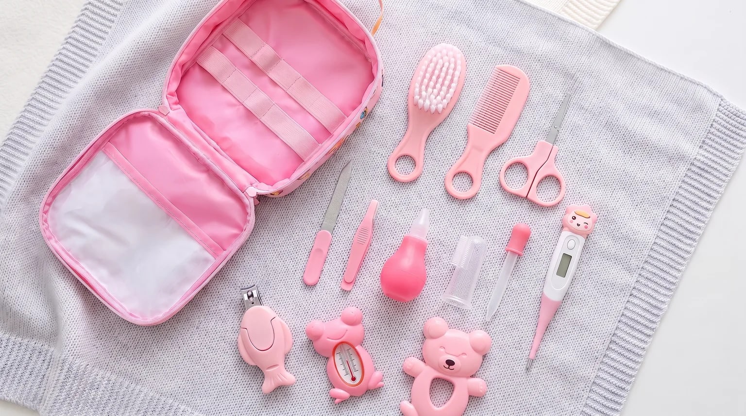 Bolsa rosa aberta e acessórios de cuidados do bebê sobre uma manta cinza, com escova, pente, tesoura, termômetro digital e termômetro de banho em formato de sapo.