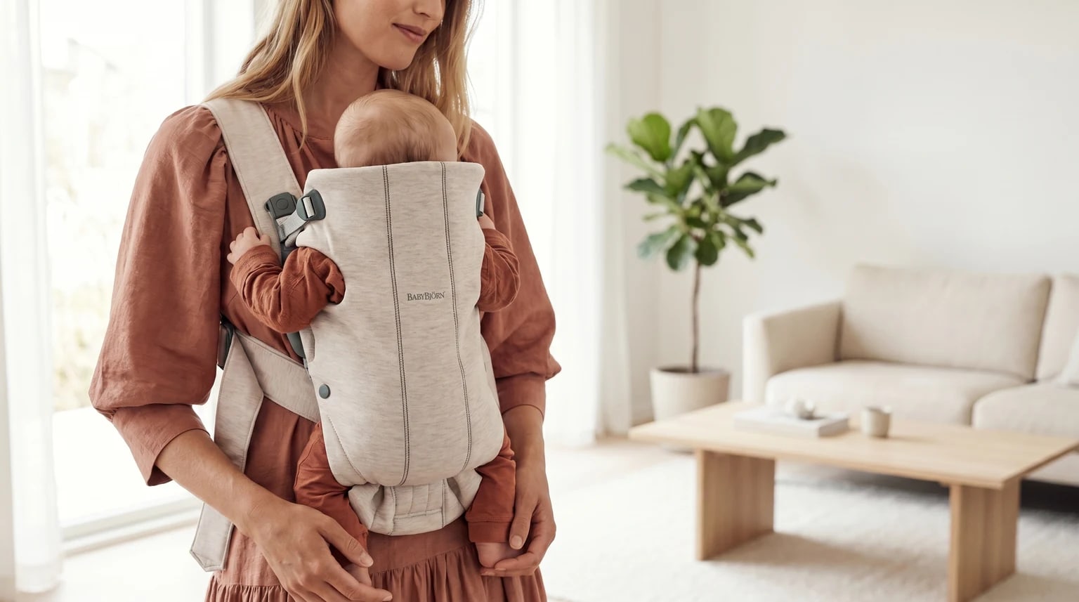 A woman carries her baby close to her chest in a light-beige BabyBjörn baby carrier, standing in a modern living room.