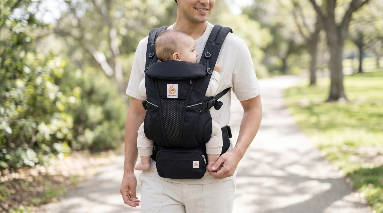 A father walks on a path through a leafy park, carrying his baby on his chest in a black Ergobaby baby carrier.