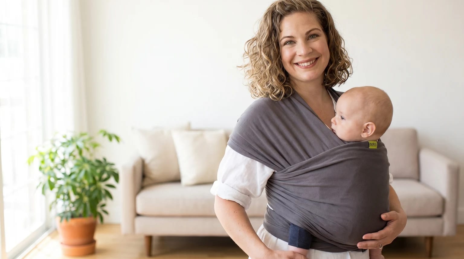 A smiling mum with curly hair holds her baby, who is snuggled close to her chest in a charcoal grey wrap carrier.