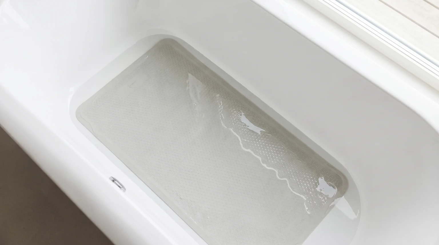 A light grey, textured bath mat lying at the bottom of a clean white bath, with a shallow layer of water over it.