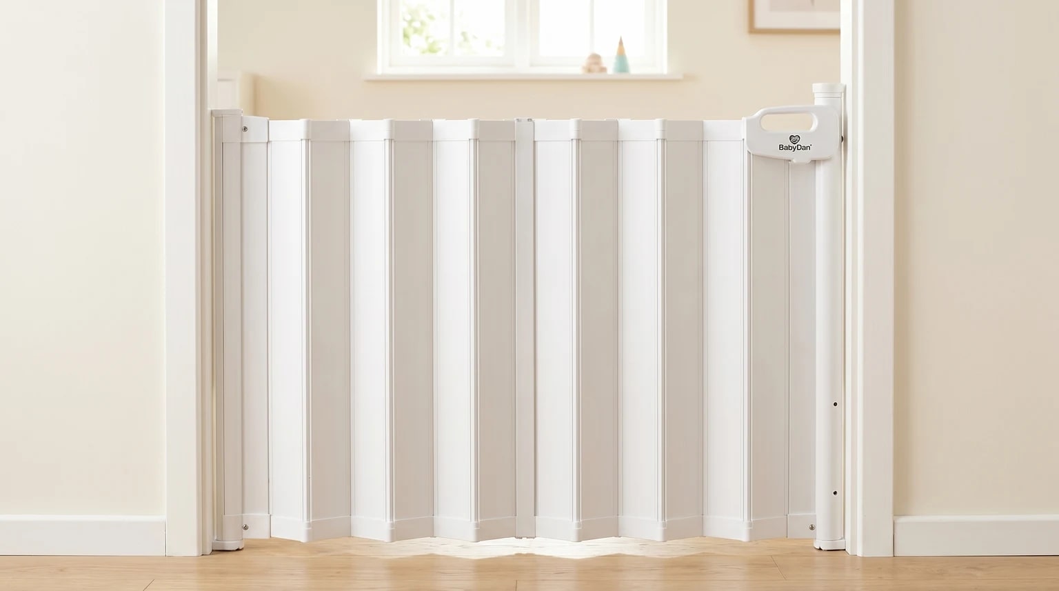 A white BabyDan Guard Me concertina safety gate installed across a bright doorway with a tidy room beyond it.