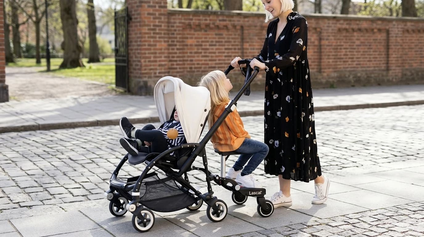A child sits on a saddle-seat buggy board attached behind a grey pushchair on a sunny pavement.