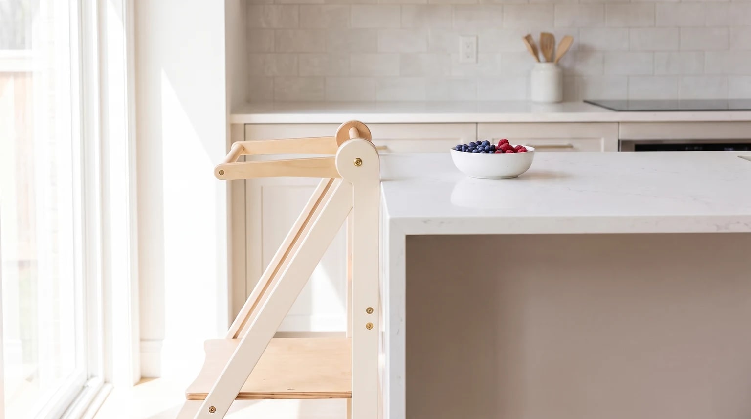 A wooden learning tower with a white finish, standing by a kitchen island with a bowl of fresh berries on the white worktop.