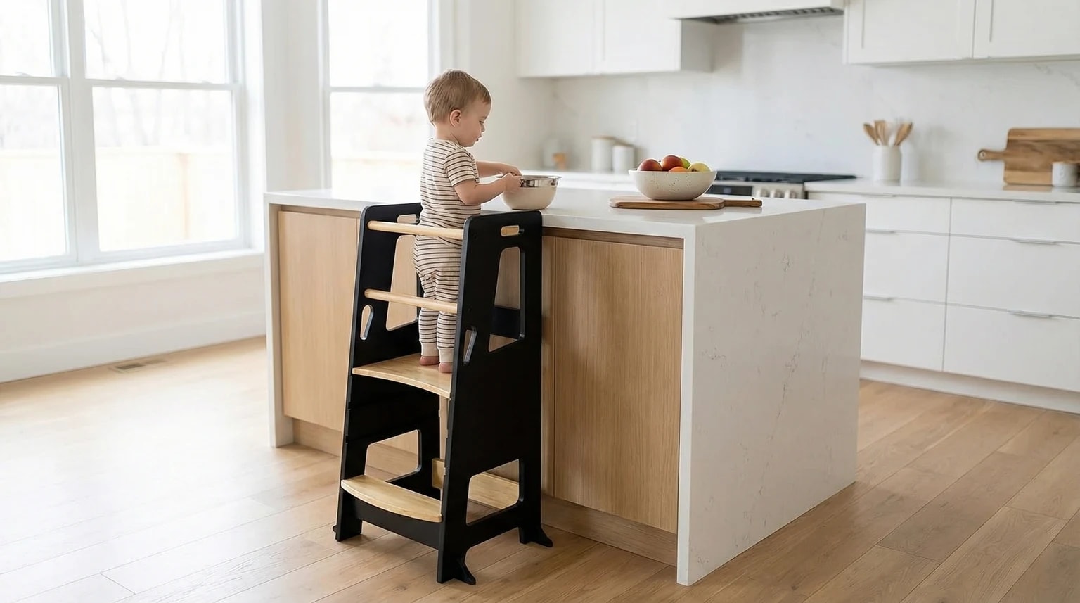 Criança pequena numa torre de aprendizagem preta e em madeira, a mexer numa taça ao balcão de uma cozinha moderna e luminosa.