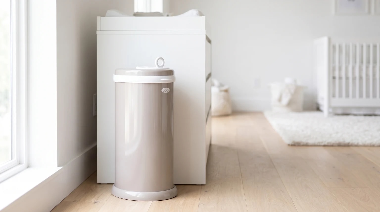 A taupe-coloured Ubbi nappy bin standing on a light wooden floor next to a white changing unit in a bright nursery.