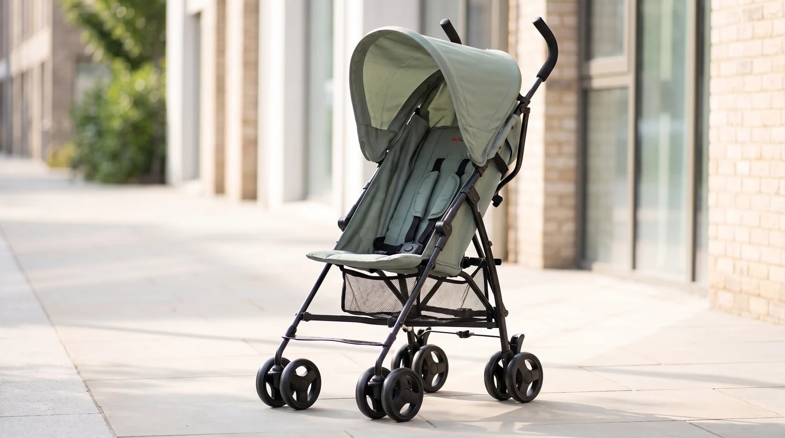 A sage green lightweight pushchair with a black frame and sun hood, standing empty on a sunlit city pavement.