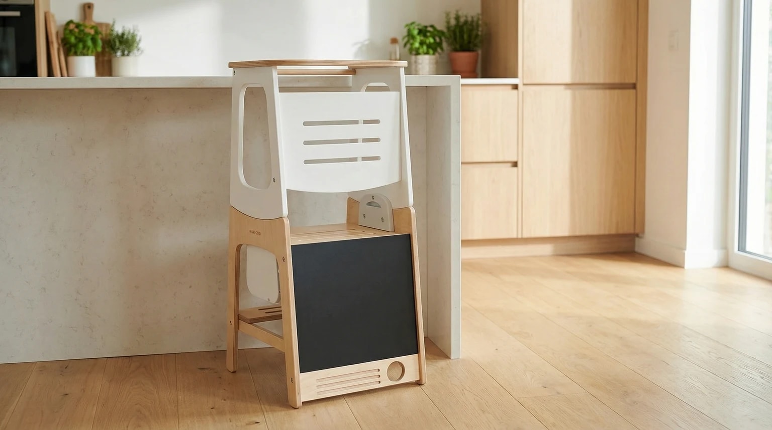 Torre de aprendizagem Maxi-Cosi em madeira clara e branca, de pé ao lado de uma bancada de cozinha moderna e bem iluminada.