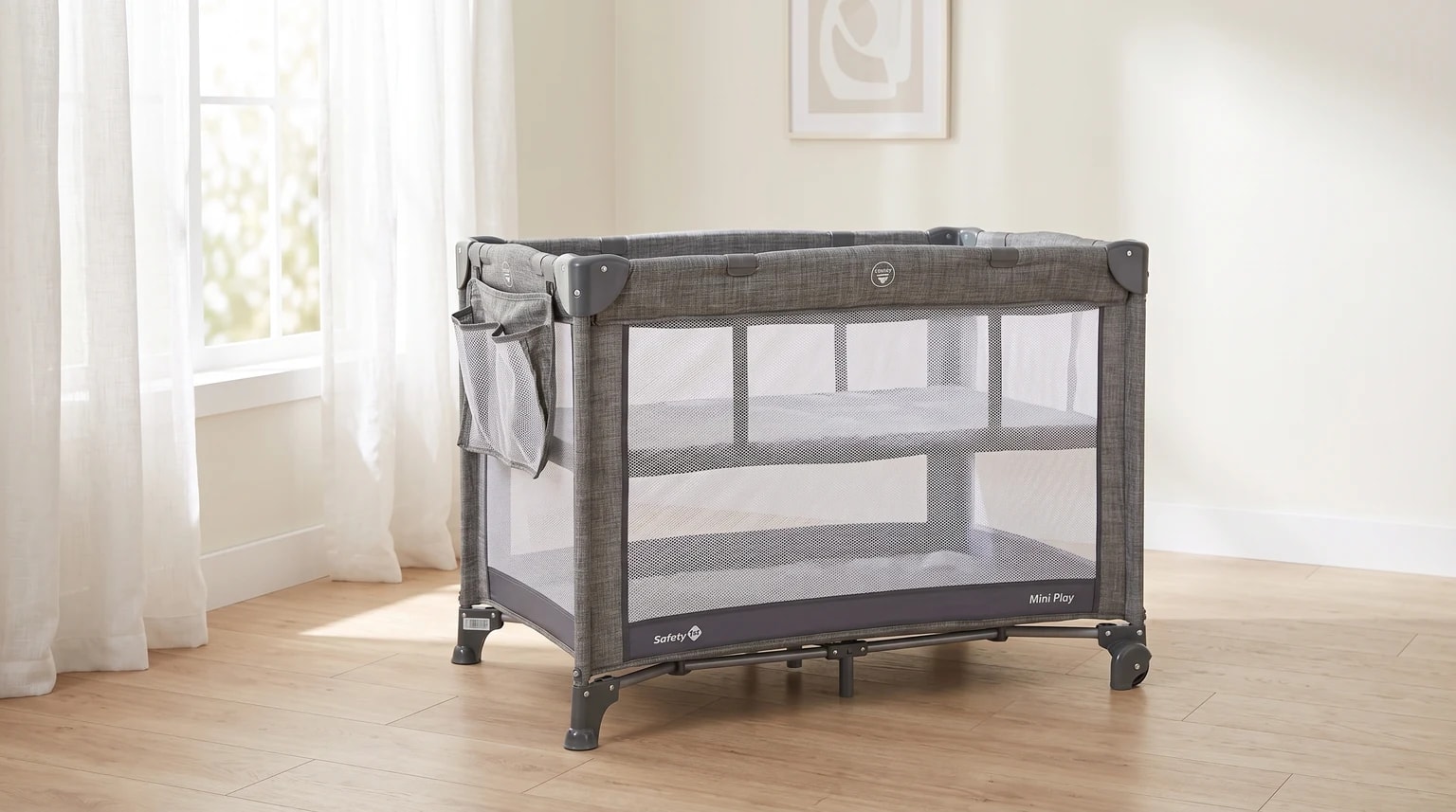 Berço portátil cinza da Safety 1st montado em um quarto com piso de madeira, ao lado de uma janela com cortinas brancas.