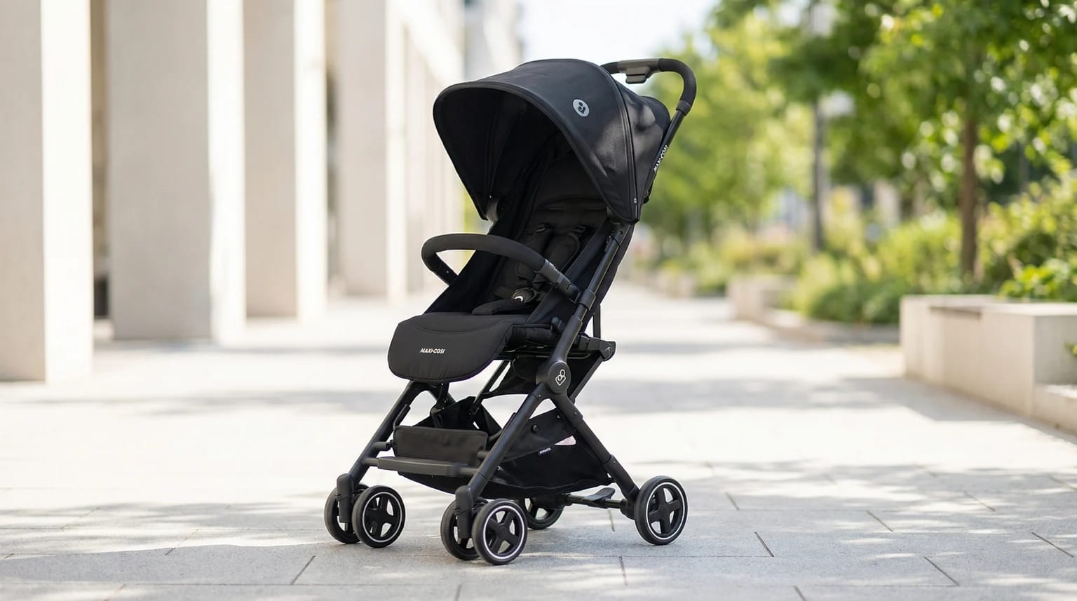 Carrinho de bebé compacto Maxi-Cosi Lara2 em preto, estacionado numa calçada moderna e soalheira de uma cidade.