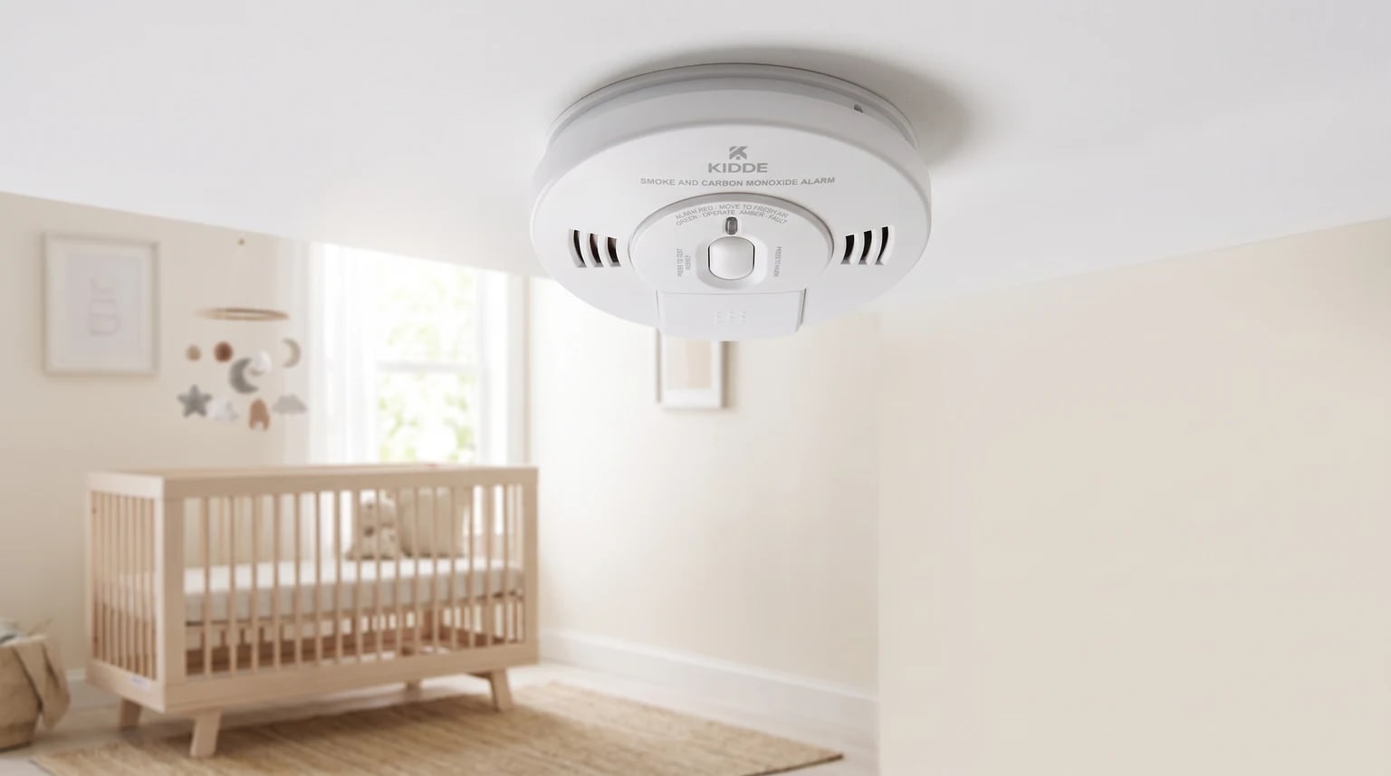 A Kidde smoke and carbon monoxide alarm fitted to the ceiling of a nursery, with a wooden cot visible in the background.