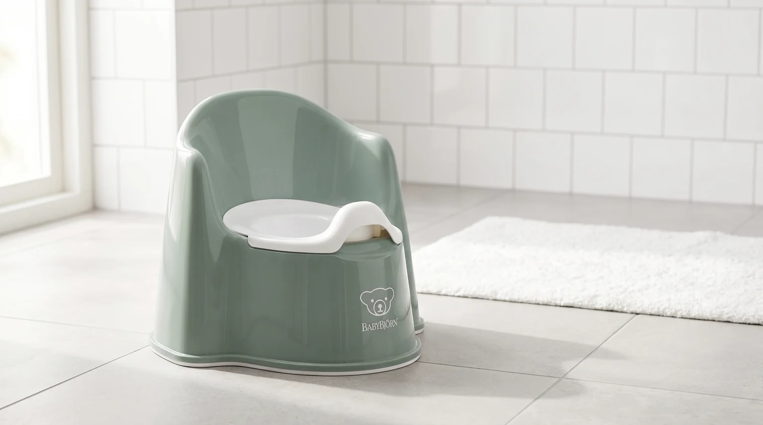 Bacio ergonómico BabyBjörn em verde-sálvia, colocado no chão de uma casa de banho moderna e luminosa.