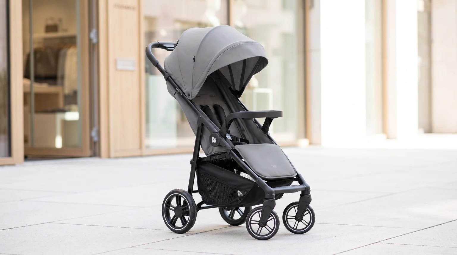 A modern grey pushchair with a black frame and large sun canopy, pictured on a pavement in front of a shop.