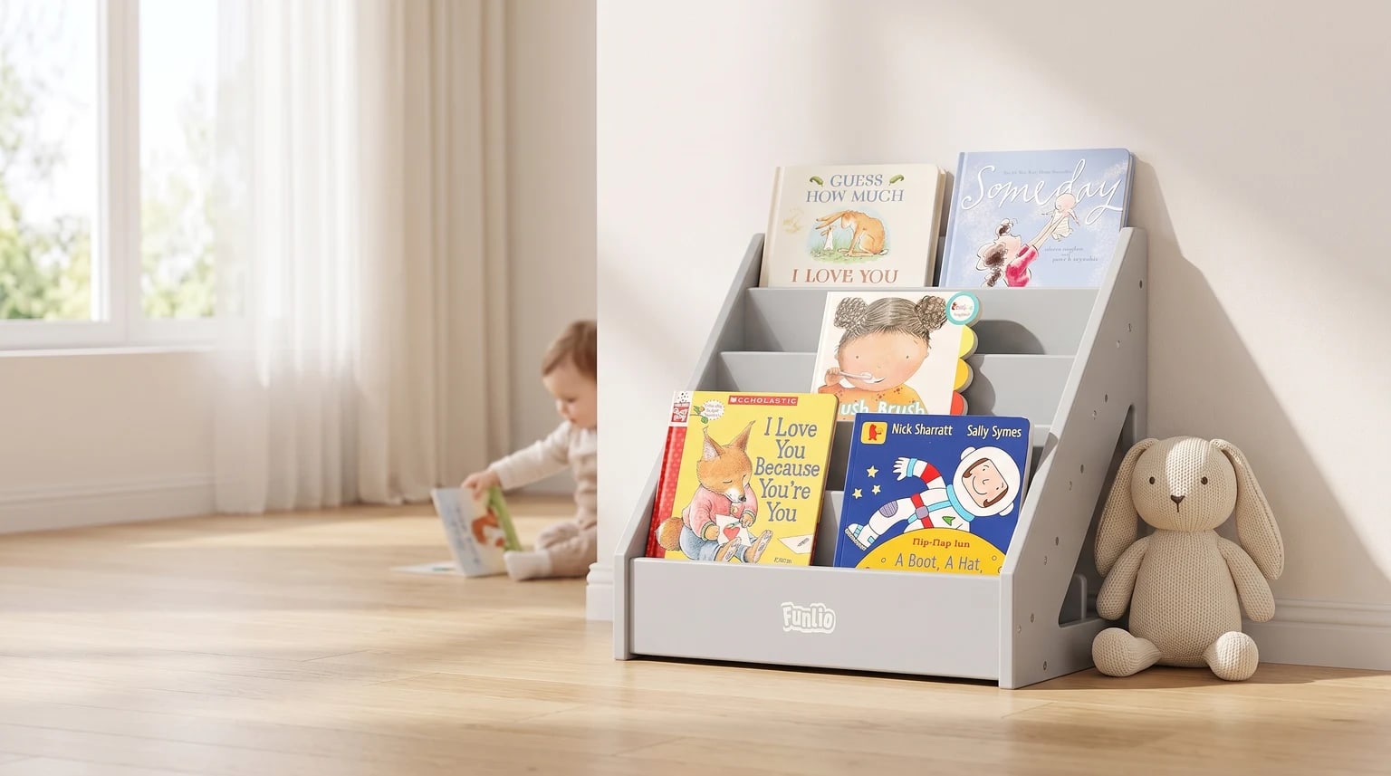 A grey, front-facing children's bookcase on a wooden floor, filled with picture books. A knitted rabbit toy sits beside it and a baby reads in the background.