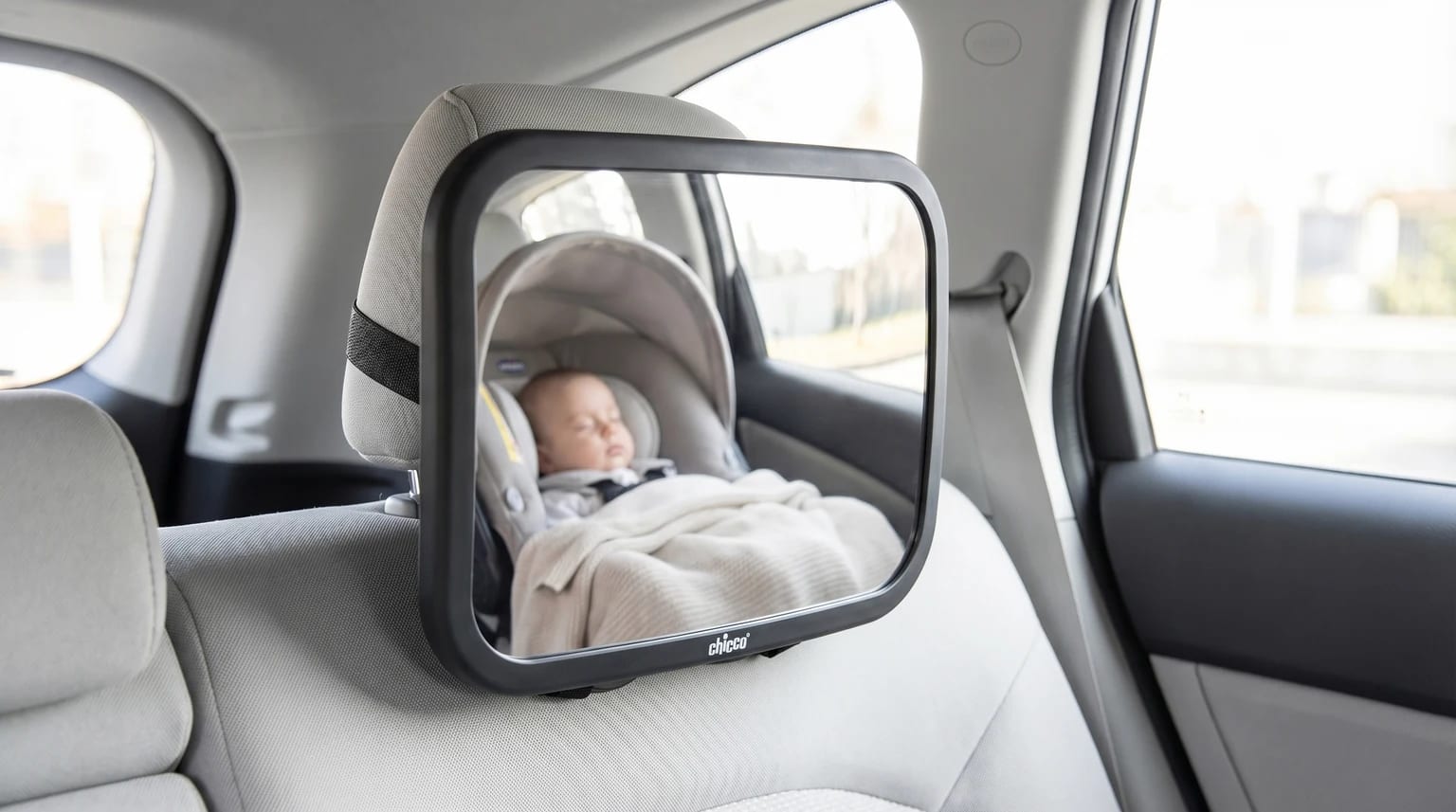 Espelho retrovisor Chicco para automóvel, montado no encosto de cabeça do banco traseiro, a refletir a imagem de um bebé a dormir pacificamente na sua cadeirinha.
