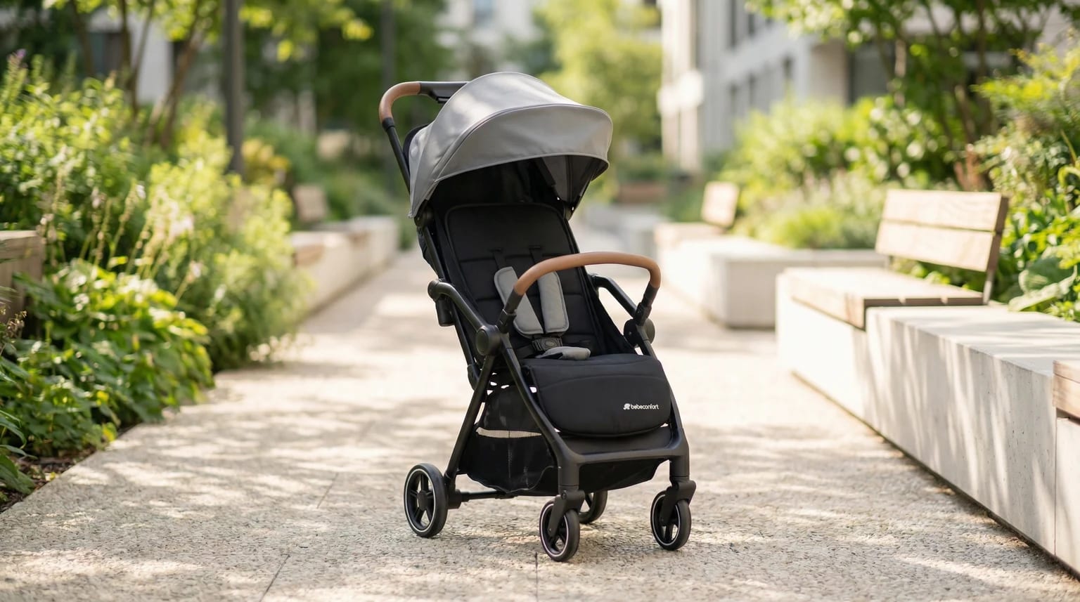 Carrinho de bebé Bebeconfort Sunlite preto e cinzento, com detalhes em pele castanha, num caminho de parque soalheiro e tranquilo.