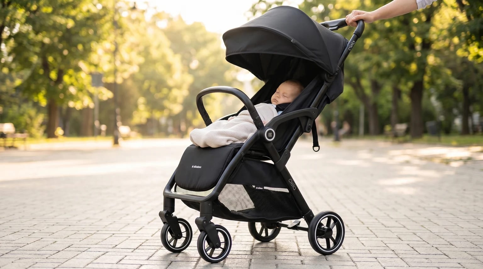 Um pai ou uma mãe empurra um carrinho de bebé KikkaBoo CIELA preto num passeio de parque luminoso e moderno.